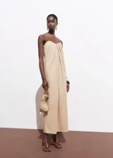 Vestido largo color beige, de diseño recto sin tirantes (palabra de honor) con escote corazón y detalle de pliegues en la parte delantera. Cuenta con cierre de cremallera en la espalda y bolsillos laterales.