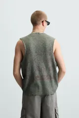 Camiseta sin mangas de punto acanalado, color verde oliva desgastado, con cuello redondo y abertura frontal en forma de 'V'. Presenta un estampado desvanecido en el centro del pecho.