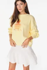 Sweater de punto color amarillo claro, con cuello redondo y diseño holgado. Cuenta con un bordado central en el pecho que representa un sol con su reflejo y la inscripción "PETRA" en tonos naranjas.