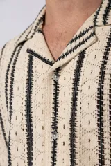 Camisa de manga corta con cuello clásico y cierre frontal de botones. Confeccionada en tejido de punto crochet calado, presenta un patrón de rayas verticales negras sobre fondo crudo.