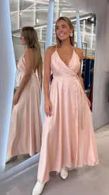 Vestido largo de satén color rosado, con diseño cruzado, escote en V pronunciado, tirantes finos tipo halter y lazo ajustable en la cintura.
