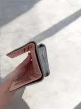 Billetera compacta de diseño plegable en color celeste, con textura granulada. Presenta una placa metálica dorada con el logo de la marca en el frente y una pequeña correa lateral del mismo tono.