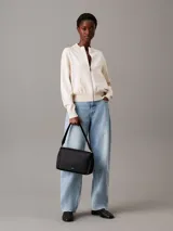 Cartera de mano Calvin Klein con correa ajustable para el hombro. El material exterior es de piel sintética negra con un patrón texturizado de monograma repetido. Presenta una solapa con cierre magnético y un logo metálico plateado en la parte frontal.