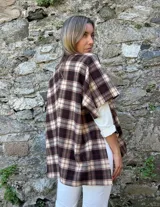 Ruana estilo chaqueta con diseño de cuadros escoceses en tonos marrones y beige. Presenta un cierre frontal tipo alamar con detalle en cuero sintético y bordes terminados.