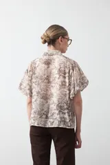 Camisa de manga corta abullonada, con cuello camisero y botones frontales. Presenta un estampado de inspiración barroca en tonos marrón y beige claro.