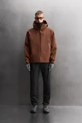 Cazadora deportiva color marrón confeccionada en tejido técnico de tres capas con membrana impermeable. Cuenta con cuello alto con capucha ajustable, mangas largas con puños reforzados ajustables, aberturas con cremallera en axilas, bolsillos frontales con cremallera y bajo ajustable con elásticos laterales.