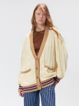 Cardigan de punto trenzado en color negro con detalles en color crema en los bolsillos, puños y ruedo. Presenta un diseño oversize, cierre frontal con botones y bordado decorativo en uno de los bolsillos delanteros.