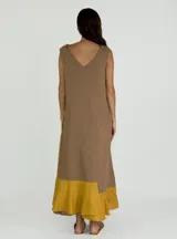 Vestido largo sin mangas de lino, con escote en V y diseño bicolor que combina un tono beige en el frente y un panel lateral en color fucsia. Presenta una costura central visible con detalle desflecado y un ruedo con volado.