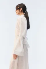 Camisa oversize negra de algodón liviano con textura calada, cuello camisero, botones frontales, mangas largas voluminosas y tiras para anudar en la espalda.