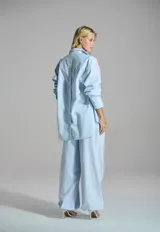 Camisa blanca de corte mullet, más larga en la parte posterior, con cuello camisero, bolsillo aplicado en el pecho, mangas largas con puños anchos y detalle de botones en la espalda.