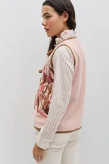 Chaleco de lana color beige con bordados florales artesanales en tonos marrones y blancos. Presenta un diseño sin mangas con volados en los hombros y cierre frontal mediante lazos negros ajustables.