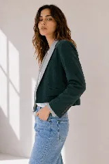 Chaqueta corta de paño en color verde petróleo, con solapas en contraste de color gris claro con textura sutil. Presenta un diseño estructurado con bolsillos frontales con solapa y cierre de un botón.