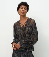 Blusa confeccionada en tul de color azul oscuro con estampado paisley en tonos beige y dorado. Cuenta con escote en V con lazo ajustable con detalles metálicos, mangas largas amplias con puños elásticos y cintura elástica que genera un leve efecto peplum. Es una prenda ligera y transparente.