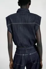 Chaleco de denim azul oscuro con cuello solapa, cierre frontal con cremallera y bajo elástico abullonado. Presenta tapetas delanteras y costuras en contraste.