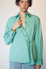 Camisa de manga larga con cuello solapa, cierre frontal con botones y diseño cruzado con lazada lateral en el mismo tejido. Presenta un estampado de rayas verticales en tonos verdes y azules.