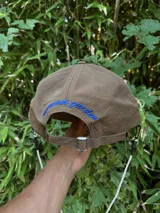 Gorra de color marrón claro con visera curva y logo bordado en azul claro en el frente.