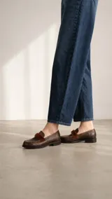 Mocasín de cuero vacuno con textura gastada en tono marrón oscuro. Presenta un detalle de banda frontal en pelo sintético al tono y costuras reforzadas en el empeine.