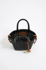 Bolso tipo cesta de estructura rígida y diseño trenzado en color negro. Cuenta con dos asas superiores cortas y una correa larga ajustable en color marrón. Incluye un dije circular decorativo con grabado en tono madera.