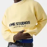 Suéter de punto grueso color amarillo, con corte cuadrado y ligeramente corto. Presenta cuello redondo, mangas largas con puños ajustados y el logo de la marca Eme Studios junto a la frase 'curated by ben ice' en jacquard oscuro en el centro del pecho.