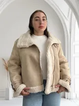 Chaqueta color beige con interior de piel sintética blanca, cierre cruzado con cremallera y detalles de hebillas en los puños.
