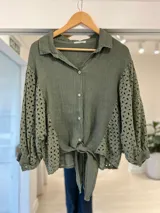 Camisa verde militar de lino, con mangas abullonadas y paneles laterales de broderie, cuello camisero, cierre frontal de botones y detalle de lazo para atar en el frente.