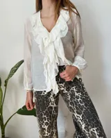 Camisa blanca traslúcida con cuello en V y volados frontales. Mangas largas con puños elásticos.