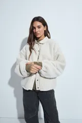 Campera de corderito color beige, con cuello camisero y cierre frontal de botones a presión. Presenta una cartera central lisa en contraste y mangas largas con puños elásticos.