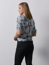 Blusa celeste con estampado blanco, de gasa, con cuello a la base y escote en V. Tiene abertura con botones, bolsillos delanteros y mangas 3/4 regulables con presilla y botón. El ruedo termina con un nudo.