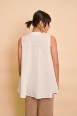Blusa sin mangas de color blanco, con cuello mao y cierre frontal de botones. Presenta un diseño holgado y asimétrico, con la parte trasera más larga y aberturas laterales en el dobladillo.