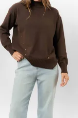 Sweater de tejido de punto color marrón, con cuello alto, corte relajado y terminaciones en rib en puños y cintura. Presenta una hilera de botones metálicos decorativos en uno de los laterales.