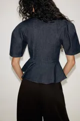 Blusa de denim oscuro con escote en pico, mangas cortas abullonadas y diseño peplum con bajo evasé. Presenta cierre frontal con alamares decorativos y cierre lateral con cremallera oculta.