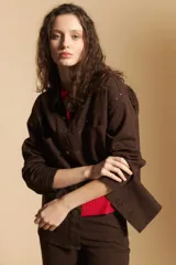Sobrecamisa de gabardina color marrón, con cierre frontal de botones, bolsillos en el pecho y detalle de tachas metálicas en los hombros y canesú. Presenta cuello clásico y mangas largas con puños abotonados.