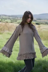 Sweater de lana merino color beige, con silueta amplia y relajada. Presenta mangas largas con volumen, terminaciones con volados de lino y flecos en los puños y en el ruedo, además de detalles trenzados a lo largo de las mangas.