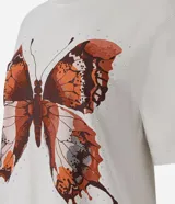 Remera oversized de algodón color gris claro, con cuello redondo y mangas cortas. Presenta una estampa frontal de mariposa marmolada en tonos tierra con pequeños detalles de strass.