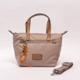 Bandolera tipo shopper color beige con diseño acolchado y pespuntes geométricos. Tiene doble asa de mano en tono tostado y una correa larga regulable y extraíble. Incluye cierre superior, bolsillo frontal con cierre adornado con un llavero de cordón naranja con cuentas y un parche decorativo.