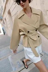Trench corto de color beige, con cuello de solapa, manga larga con hombreras y trabillas con botones. Cuenta con cierre frontal cruzado con botones oscuros y cinturón ajustable del mismo tejido.