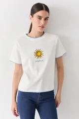 Remera blanca de algodón con cuello redondo, manga corta y bordes con detalle de costura en hilo celeste. Presenta un bordado central de un sol amarillo y la palabra 'SAVIA' debajo.