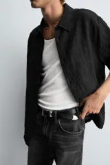 Camisa negra de manga corta con cuello solapa y textura arrugada o plisada. Corte relajado (relaxed fit) con aberturas laterales y cierre frontal de botones.