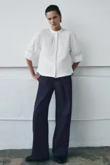 Camisa blanca de algodón con cuello redondo y manga corta abullonada. Presenta un canesú con pliegues verticales en la parte delantera y cierre frontal con botones ocultos. El bajo es redondeado.