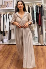 Vestido largo de seda en tono beige, con cuello camisero, botones frontales y mangas largas arremangadas. Presenta una falda con caída fluida y un cinturón trenzado marrón que marca la cintura.