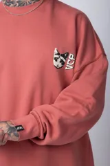 Buzo de tejido friza color crema, de corte oversize y cuello redondo. Presenta un pequeño bordado o parche en el pecho izquierdo con un diseño de cabeza de animal en tonos rojizos/terracota.