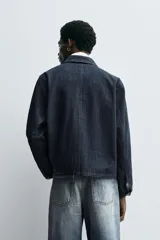 Chaqueta de denim de algodón en color azul oscuro, con cuello solapa y manga larga. Presenta cierre frontal con cremallera oculta bajo una solapa con alamares metálicos, bolsillo de vivo en el pecho y bolsillos de plastrón con solapa en la cadera.