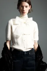 Saco de punto suave color crema, de manga corta y cuello redondo. Presenta cierre frontal con botones forrados y una moña decorativa de organza transparente en el escote. Posee terminaciones acanaladas en puños y ruedo.