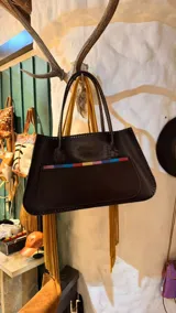 Cartera de cuero marrón oscuro, con manijas de hombro y un bolsillo frontal decorado con detalles coloridos.