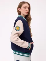 Campera estilo varsity con cuerpo en color marrón y mangas en tono beige. Presenta cierre frontal con botones a presión, cuello, puños y ruedo con franjas tejidas en verde y blanco. Incluye un parche con la letra R en el pecho y detalles bordados en las mangas.