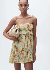 Vestido corto de gasa con estampado floral en tonos amarillos, naranjas y verdes. Presenta un diseño recto con escote cuadrado, tirantes finos dobles y un detalle de volante horizontal en la parte superior. Incluye forro interior y cierre de cremallera lateral.