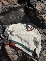 Buzo tejido de punto con escote en V, de color crema con detalles de rayas en tonos verde y bordó en el cuello, puños y ruedo. Presenta un diseño acanalado y logo bordado en el pecho.