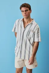 Camisa de manga corta a rayas verticales en tonos azul y blanco, con cuello camisero, cierre frontal de botones y un bolsillo en el pecho con etiqueta aplicada.