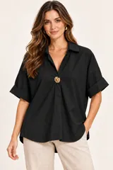 Camisa de algodón negra de corte recto y holgado, con cuello camisero, escote en V pronunciado decorado con un gran botón dorado central, mangas cortas con vuelta y dobladillo asimétrico más largo en la parte trasera.
