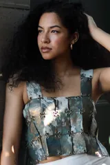 Top corto estilo corsé o armadura, compuesto por paneles rectangulares de material con estampado abstracto en tonos verde azulado, gris y dorado/bronce, unidos entre sí mediante ojales y ganchos metálicos. Tiene tirantes anchos.